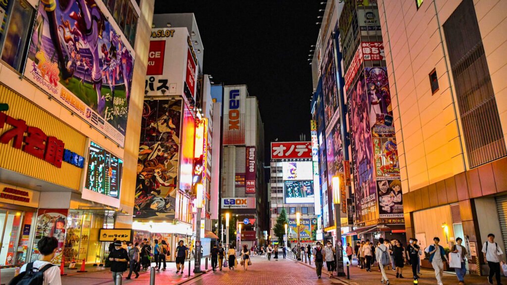 Bustling nighttime street view of Akihabara, Tokyo, Japan, with bright neon signs, anime and gaming billboards, and people walking through the vibrant shopping district known for its electronics, manga, and pop culture stores.