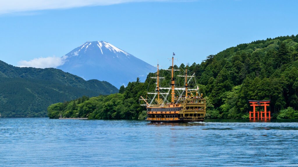 Scenic view of Lake Ashi in Hakone, Japan, with a pirate-style sightseeing cruise sailing across the water, the red torii gate of Hakone Shrine on the shoreline, and Mount Fuji towering in the background on a clear day.