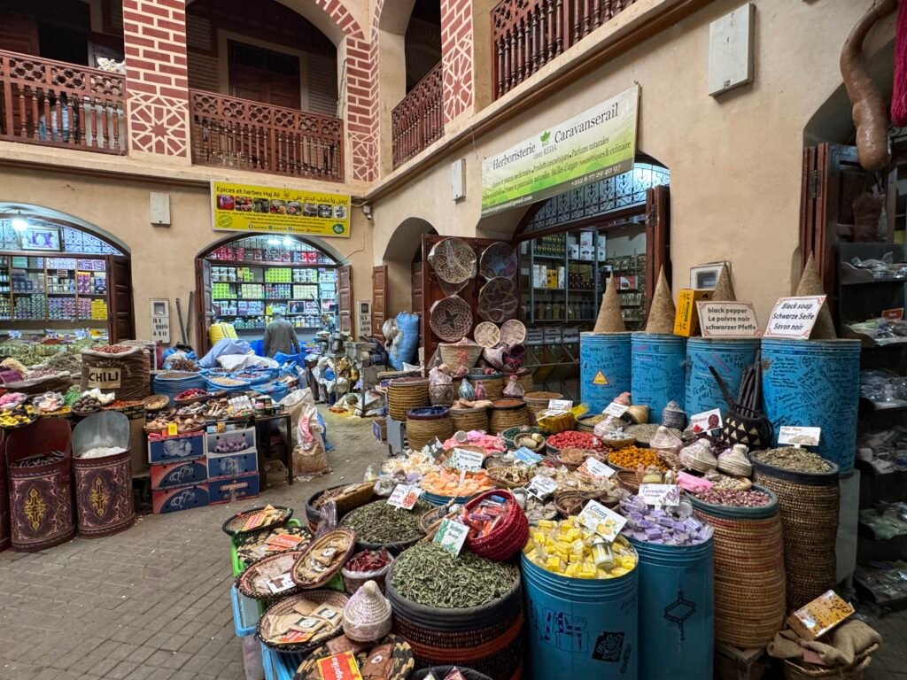 A vibrant spice shop in the Marrakech souks, displaying baskets of colorful spices, herbs, and traditional Moroccan products in a bustling market setting.