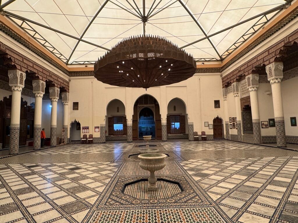 Interior of the Marrakech Museum, showcasing traditional Moroccan architecture with intricate tilework, carved arches, and a grand chandelier.