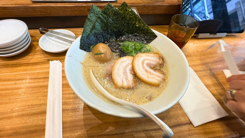 A delicious bowl of tonkotsu ramen served at a ramen shop in Tokyo, featuring rich pork bone broth, chashu pork slices, marinated soft-boiled egg, nori seaweed, and green onions, presented on a wooden counter with dining utensils.