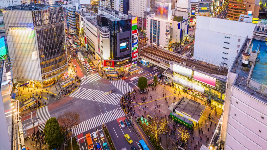 Aerial view of Shibuya Crossing in Tokyo, Japan, at dusk, showcasing the bustling intersection with pedestrians crossing in multiple directions, surrounded by neon-lit buildings, busy streets, and a vibrant cityscape.