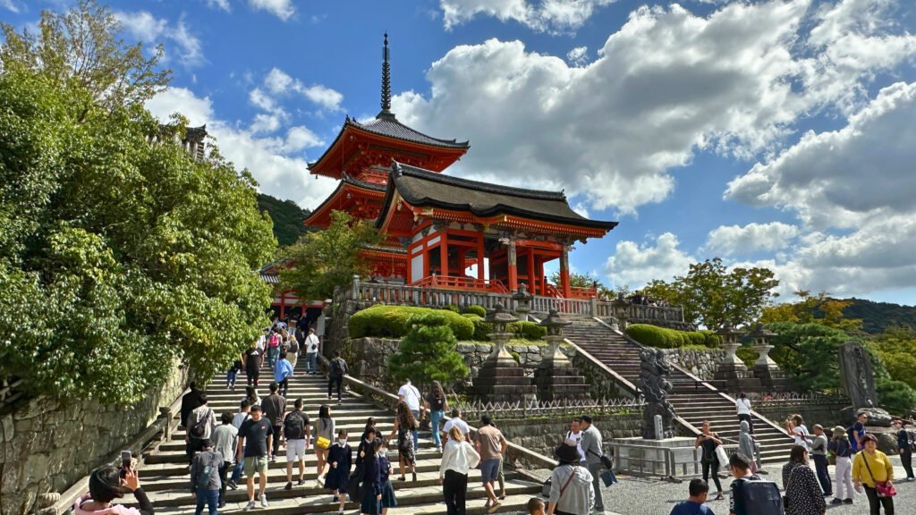 Tourists walking up the stone steps leading to Kiyomizu-dera Temple in Kyoto, Japan. The vibrant red pagoda stands against a backdrop of a bright blue sky with scattered clouds, surrounded by lush greenery. One of Kyoto’s most famous landmarks, this UNESCO World Heritage site offers breathtaking views and rich cultural history.