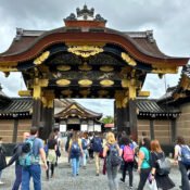 A group of tourists walking towards the elaborately decorated Karamon Gate at Nijo Castle in Kyoto, Japan. The gate features intricate wood carvings and gold embellishments, showcasing traditional Japanese architectural elegance. The cloudy sky and historic surroundings add to the grandeur of this UNESCO World Heritage site.