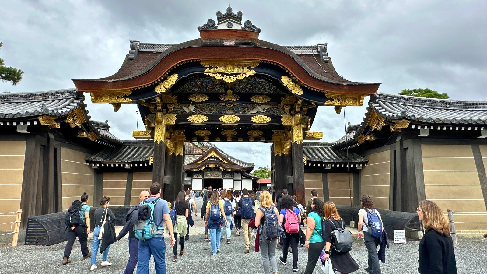 A group of tourists walking towards the elaborately decorated Karamon Gate at Nijo Castle in Kyoto, Japan. The gate features intricate wood carvings and gold embellishments, showcasing traditional Japanese architectural elegance. The cloudy sky and historic surroundings add to the grandeur of this UNESCO World Heritage site.